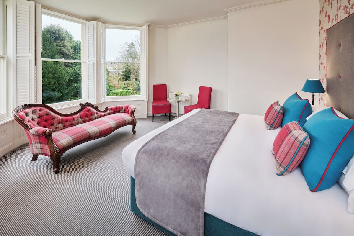 A bedroom with a large bed, a red armchair, a small table, and a window with a view of greenery.