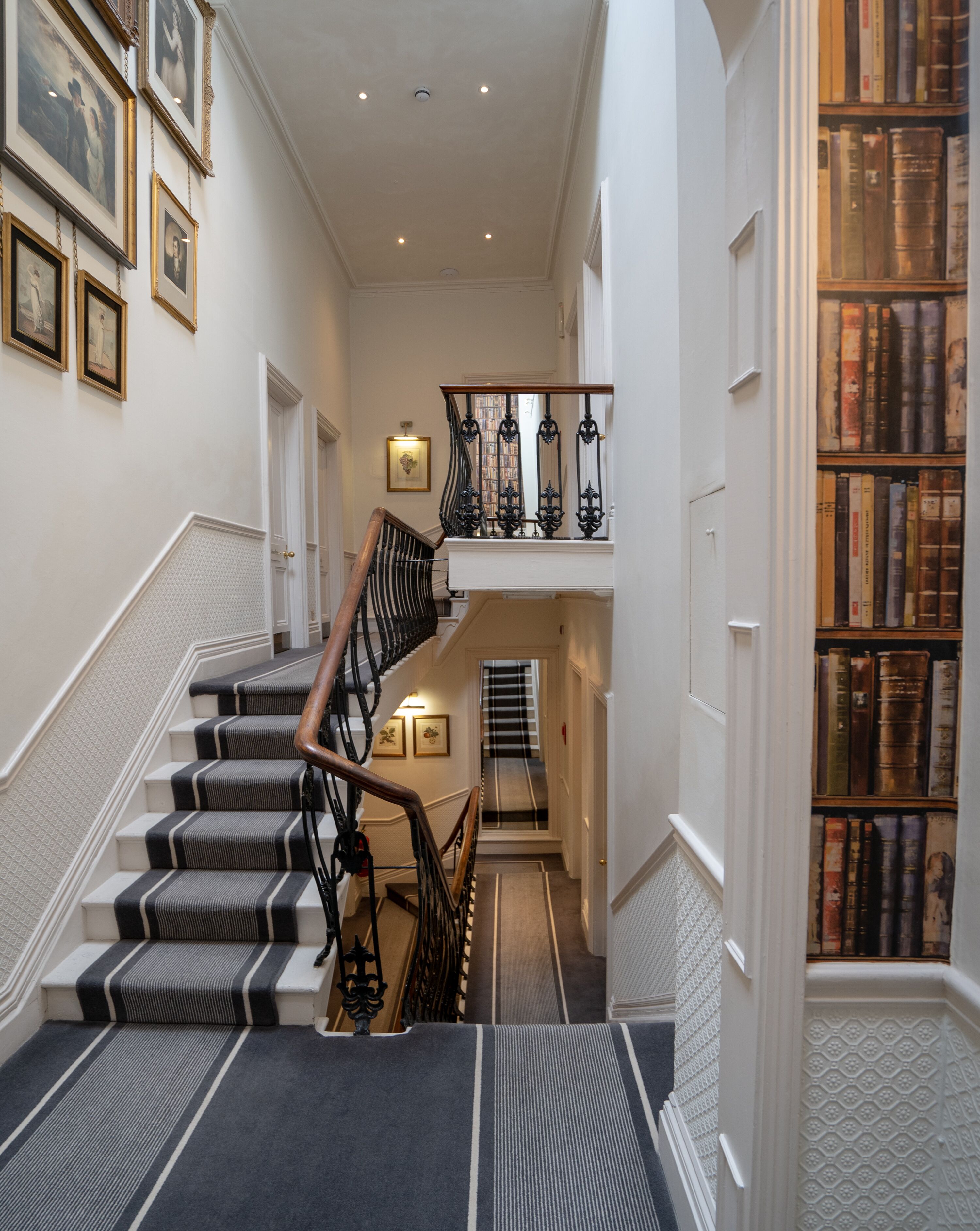 A grand staircase with a black and white striped carpet, a wooden handrail, and a bookcase filled with vintage books.