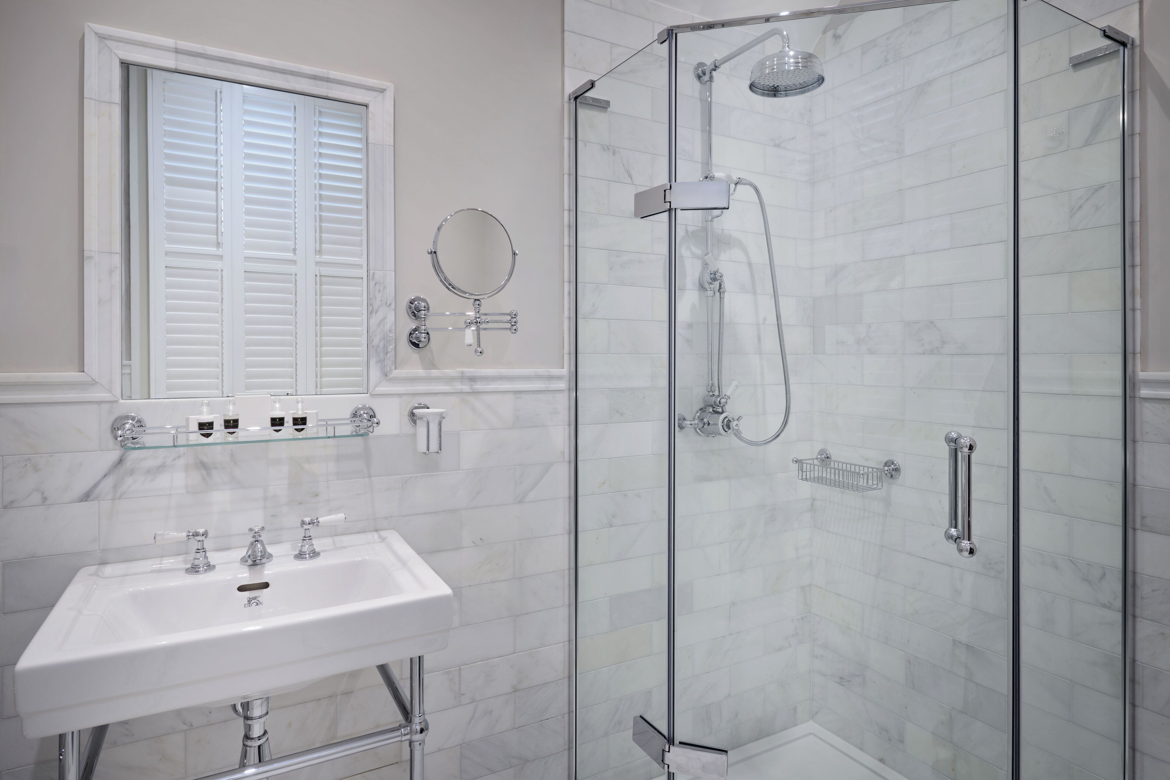 A bathroom with a glass-enclosed shower, a white sink, and a window with white shutters.