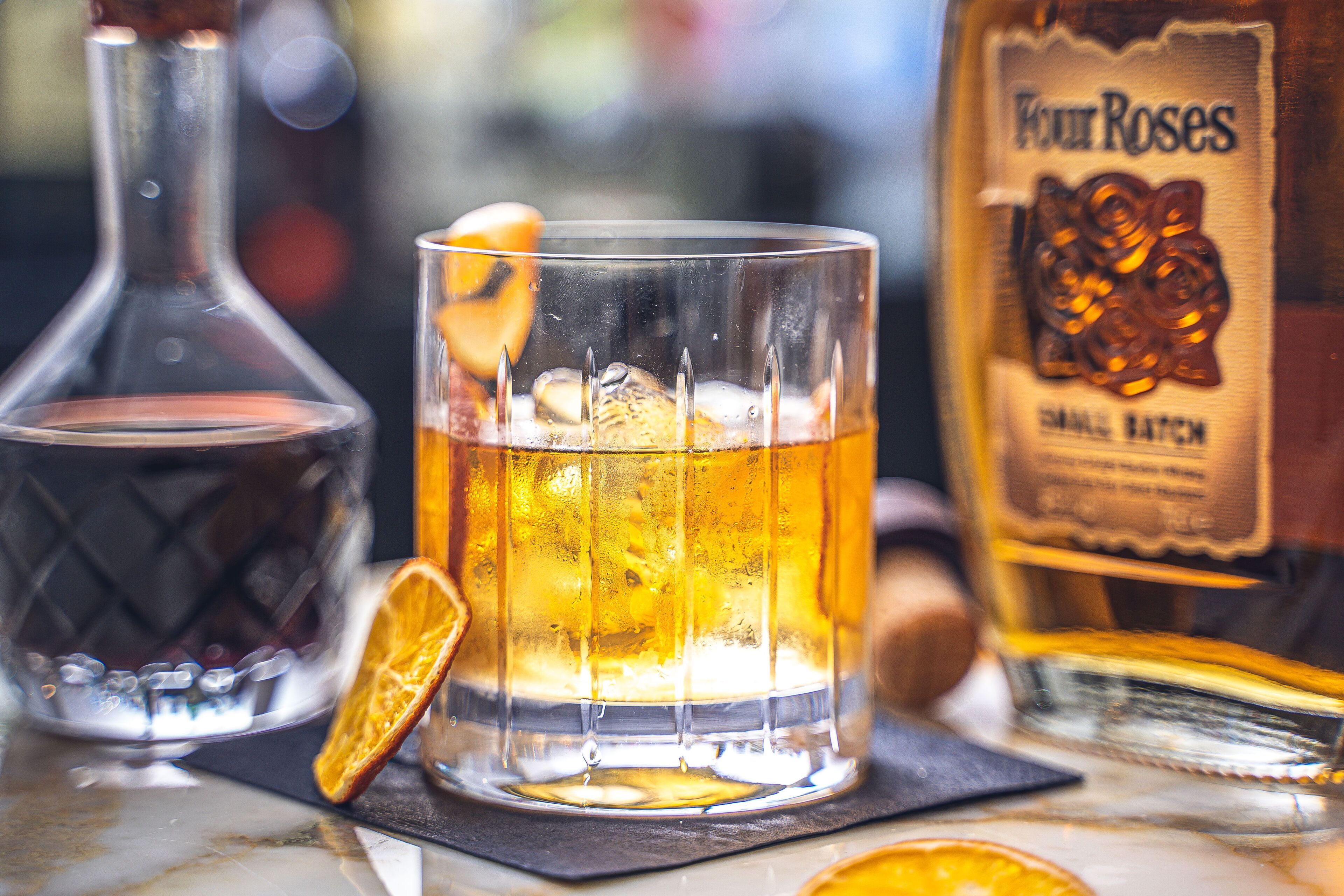 A glass of amber liquid with ice cubes and a slice of orange, a bottle of Four Roses whiskey, and a decanter.