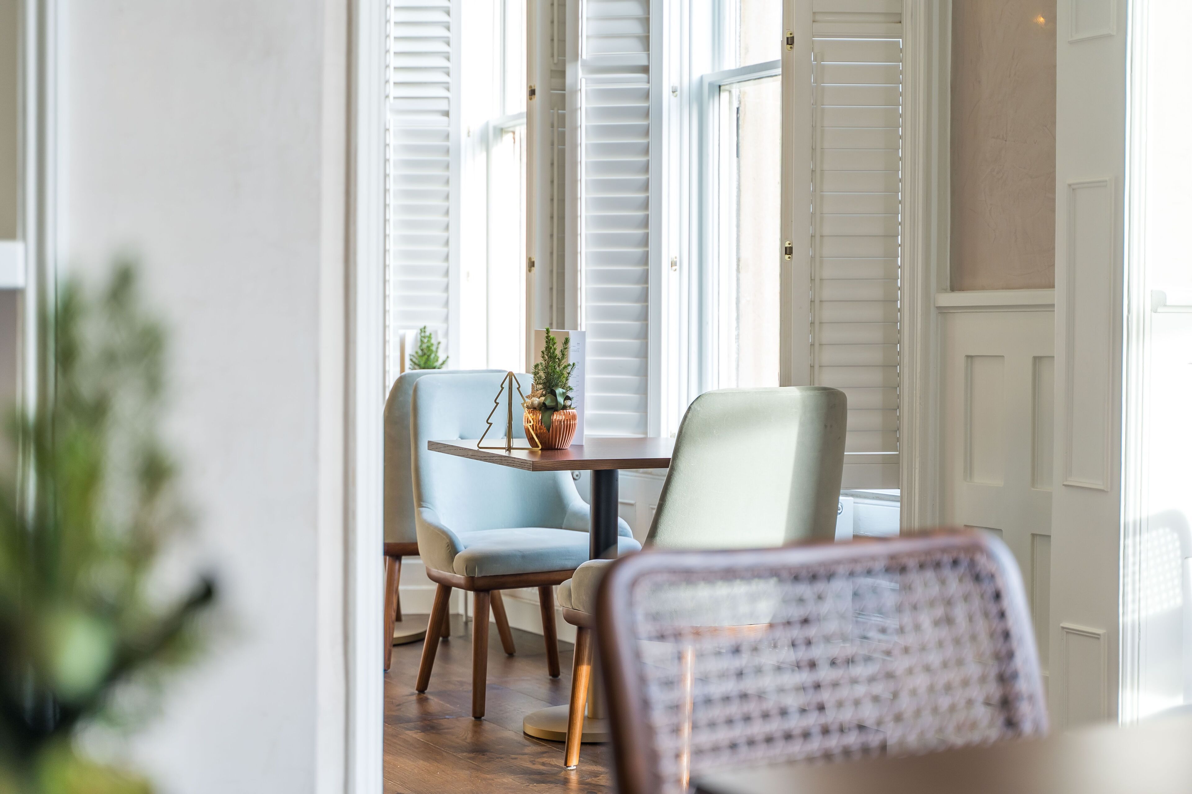 Un coin repas comprenant une table et deux chaises, une plante en pot et une vue sur une porte avec des volets blancs.