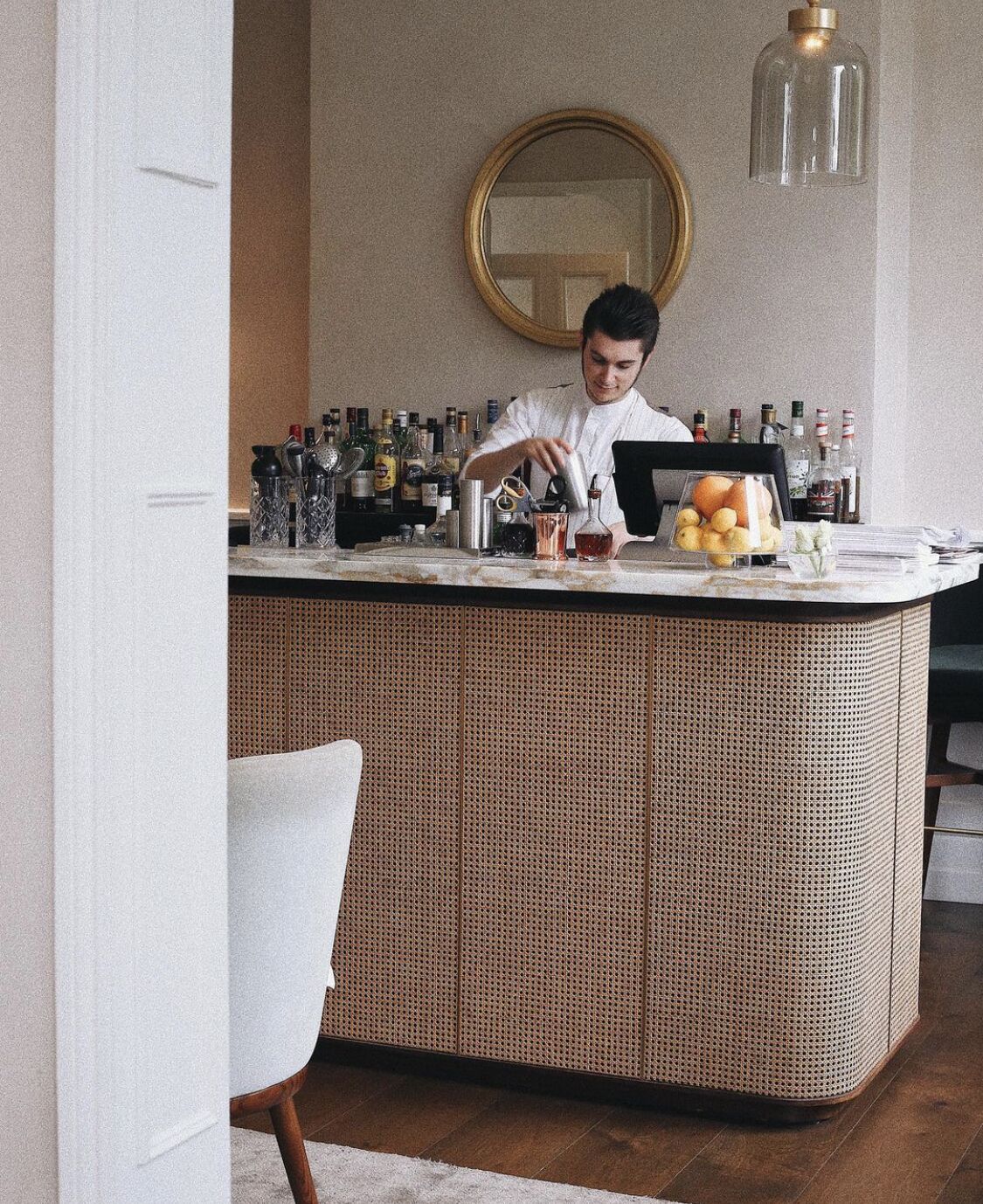 A bar area with a bartender preparing drinks, a mirror, and various bottles and fruits on the counter.