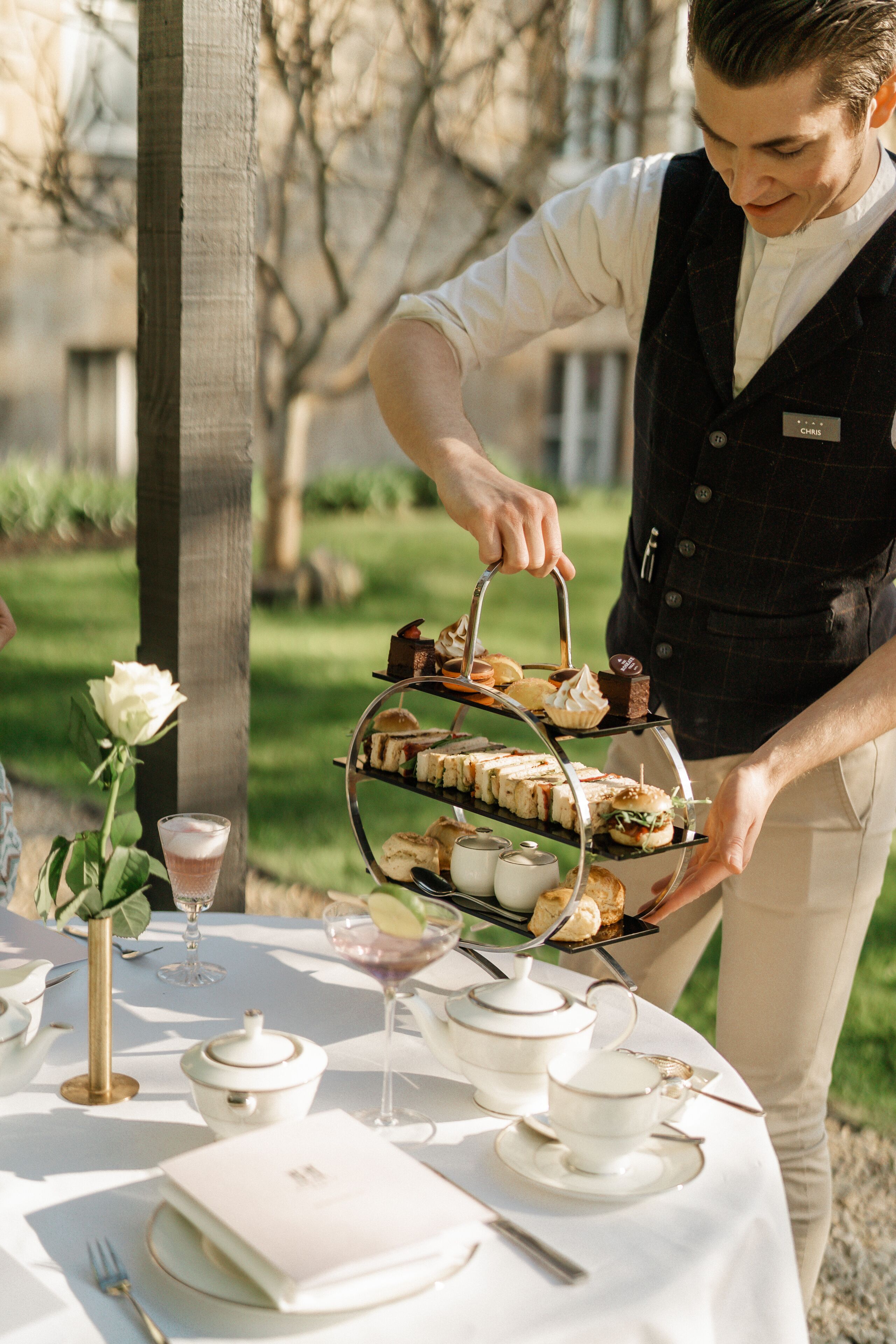 A table set for a tea service with a tiered tray of sandwiches and pastries, a teapot, cups, and a rose.