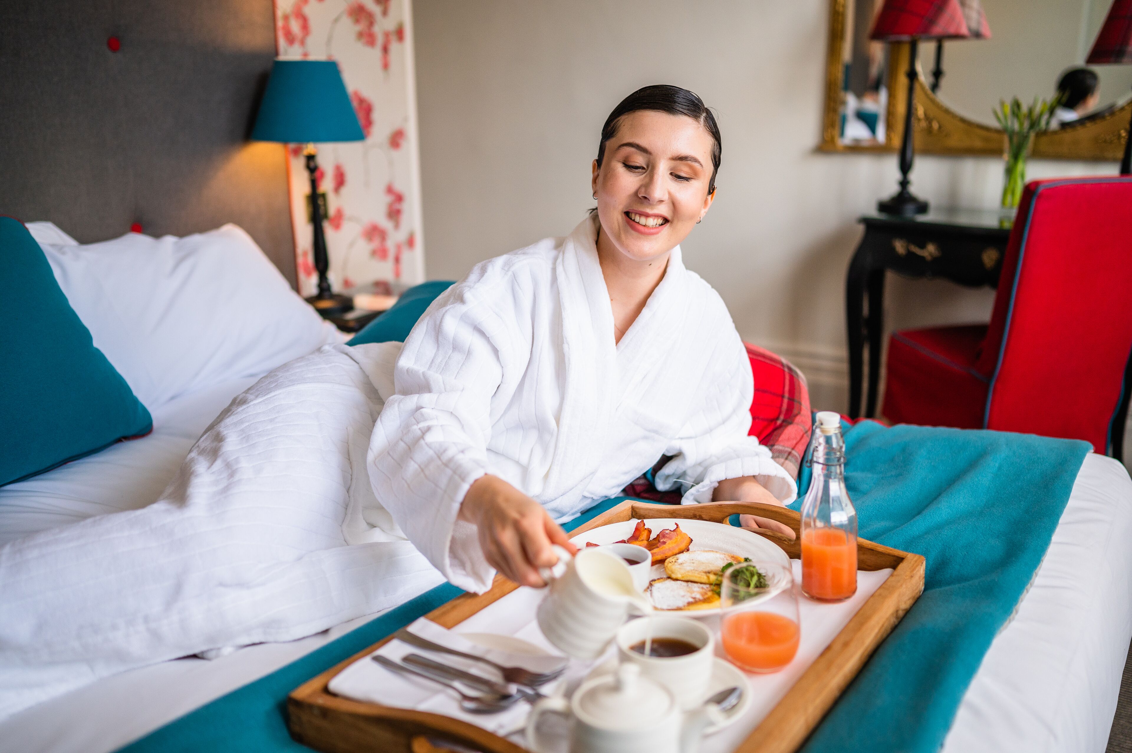 A hotel room with a bed, a tray with breakfast items, and a lamp in the background.