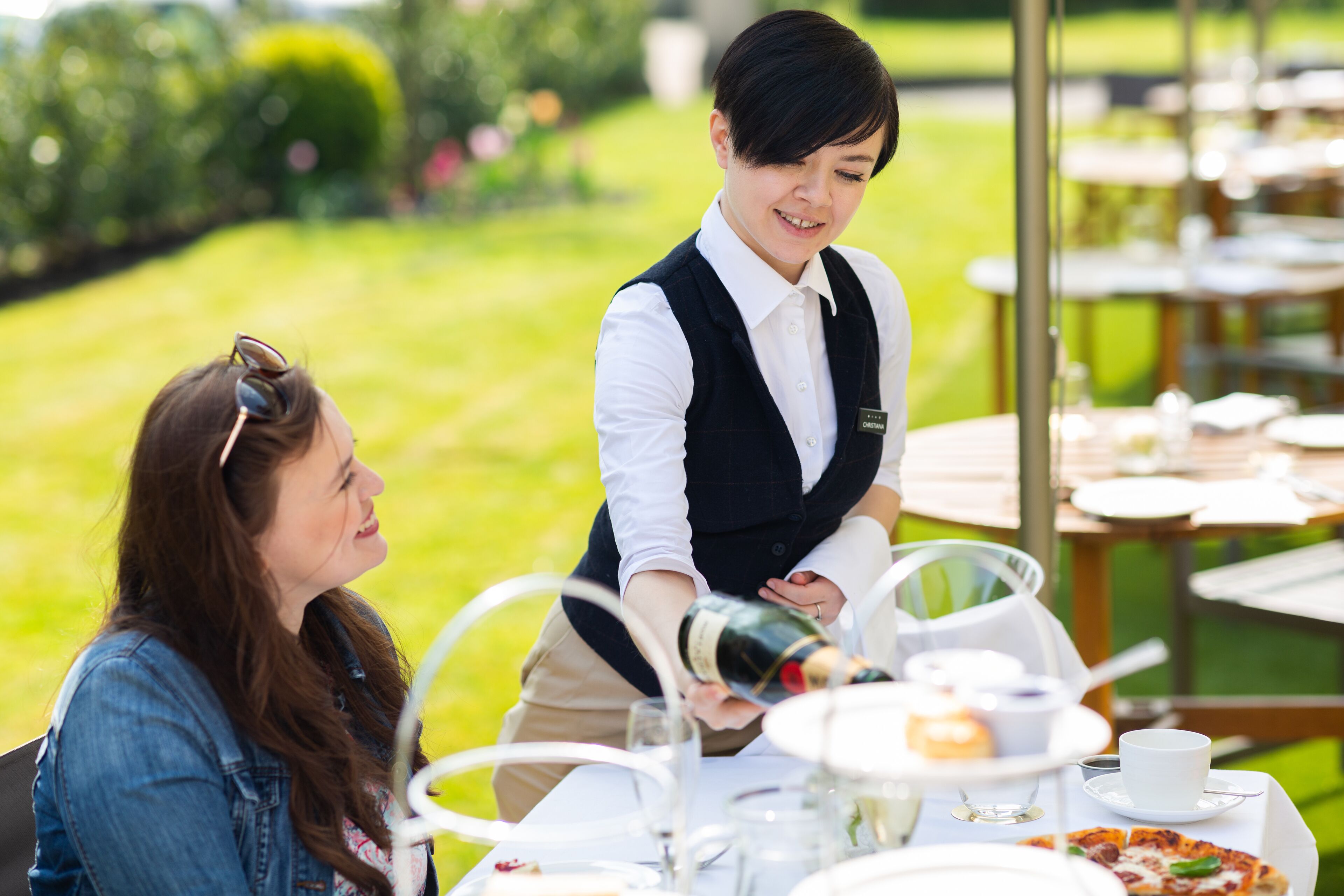 A waiter pouring a drink at an outdoor dining table with a garden view.