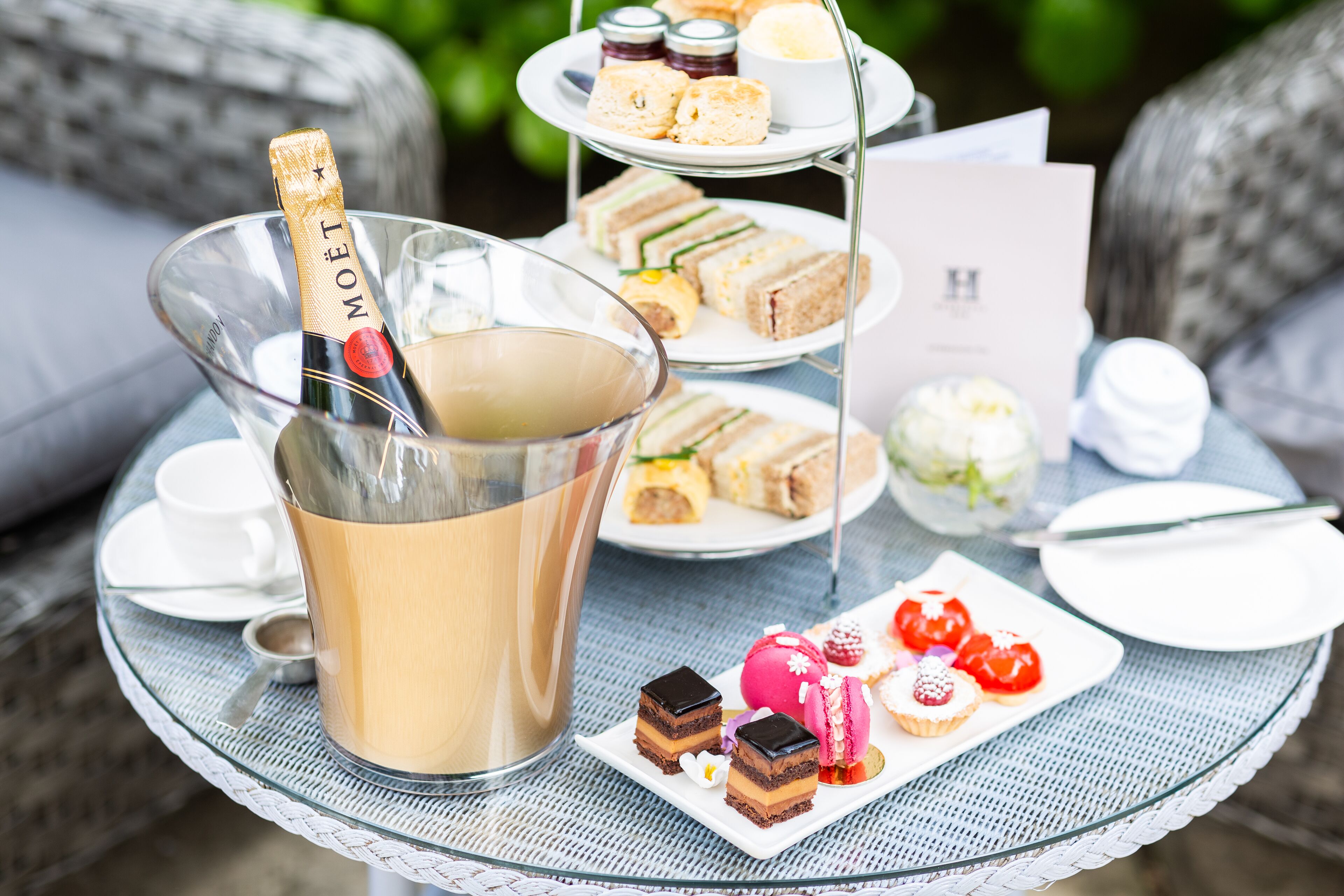 A glass table with a bottle of champagne, a tiered dessert stand, and various pastries.
