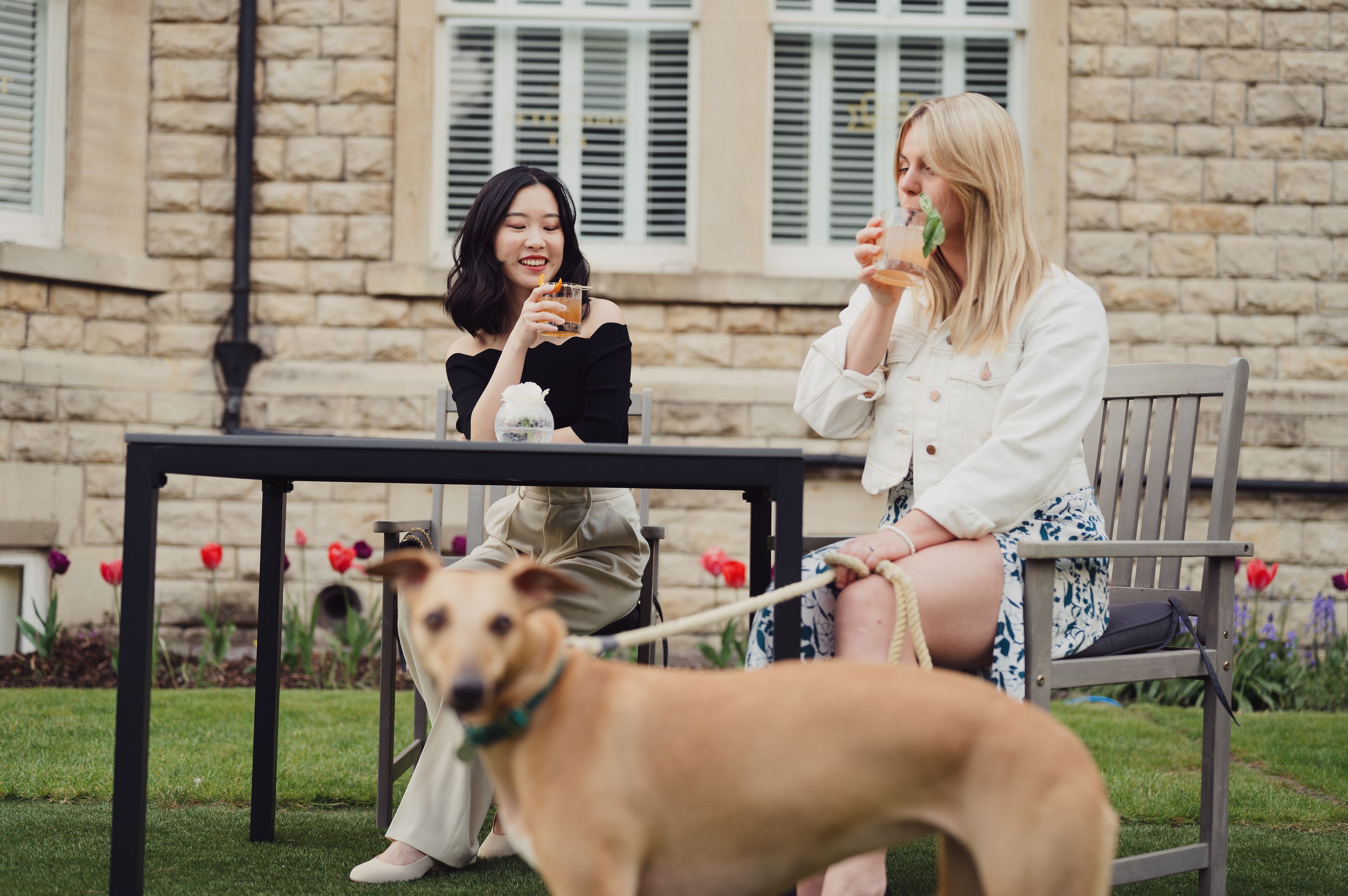 Deux personnes sont assises à une table à l’extérieur, et un chien est tenu en laisse à côté d’elles.