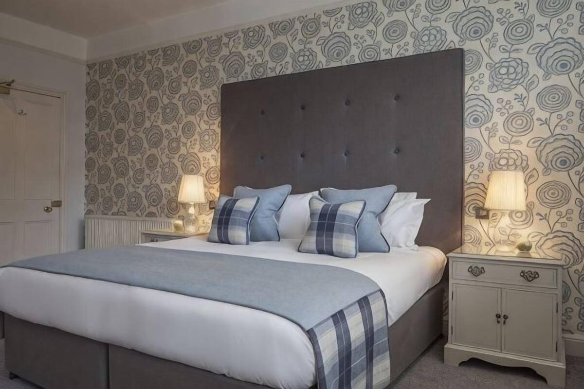 A bedroom with a large bed, two bedside tables with lamps, patterned wallpaper, and a dark headboard.
