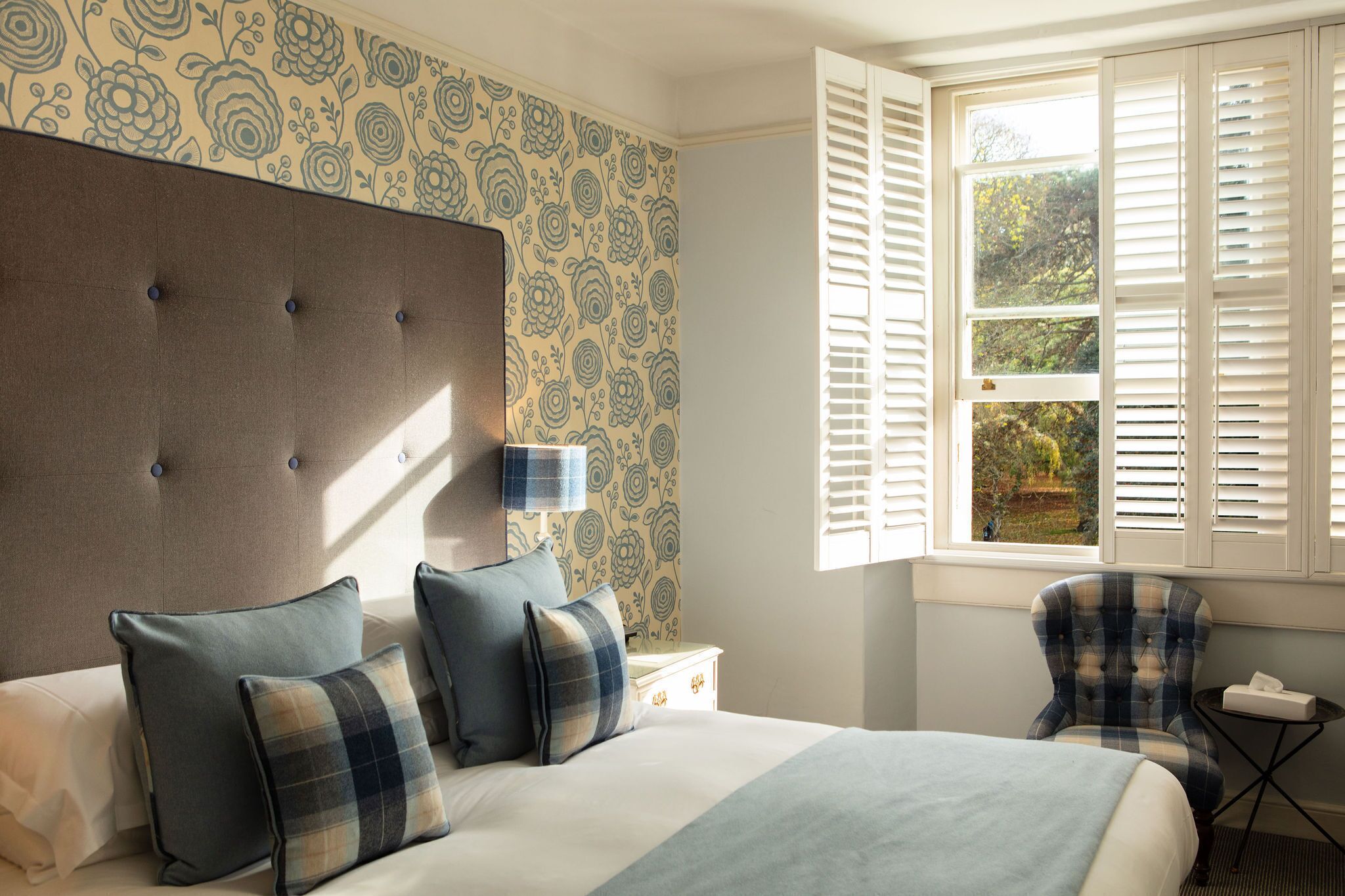 A bedroom with a tufted headboard, a bed with plaid bedding, a nightstand, a chair, and a window with shutters.