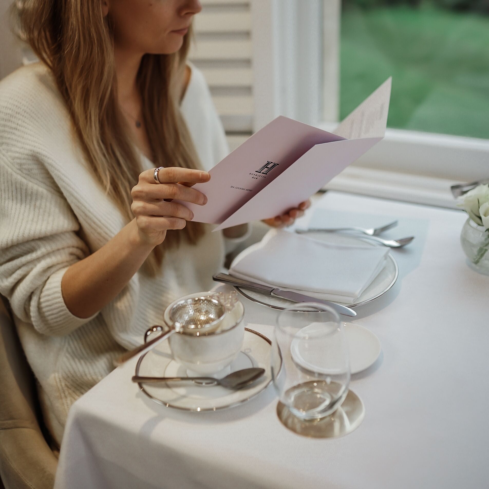 A person reading a menu at a table with a white tablecloth, a teapot, and a cup.