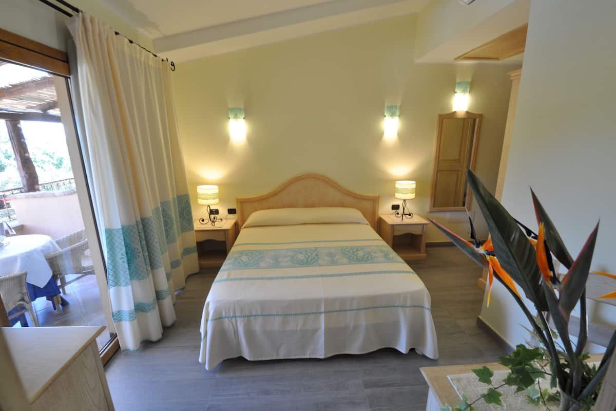 Ein Hotelzimmer mit einem Bett, zwei Nachttischen mit Lampen, einem großen Fenster mit Vorhängen, einem Holz-Kopfteil und einer Topfpflanze.