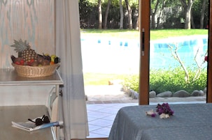 Una habitación con cama, una cesta de mimbre para frutas en un estante y vistas a una piscina y jardín.