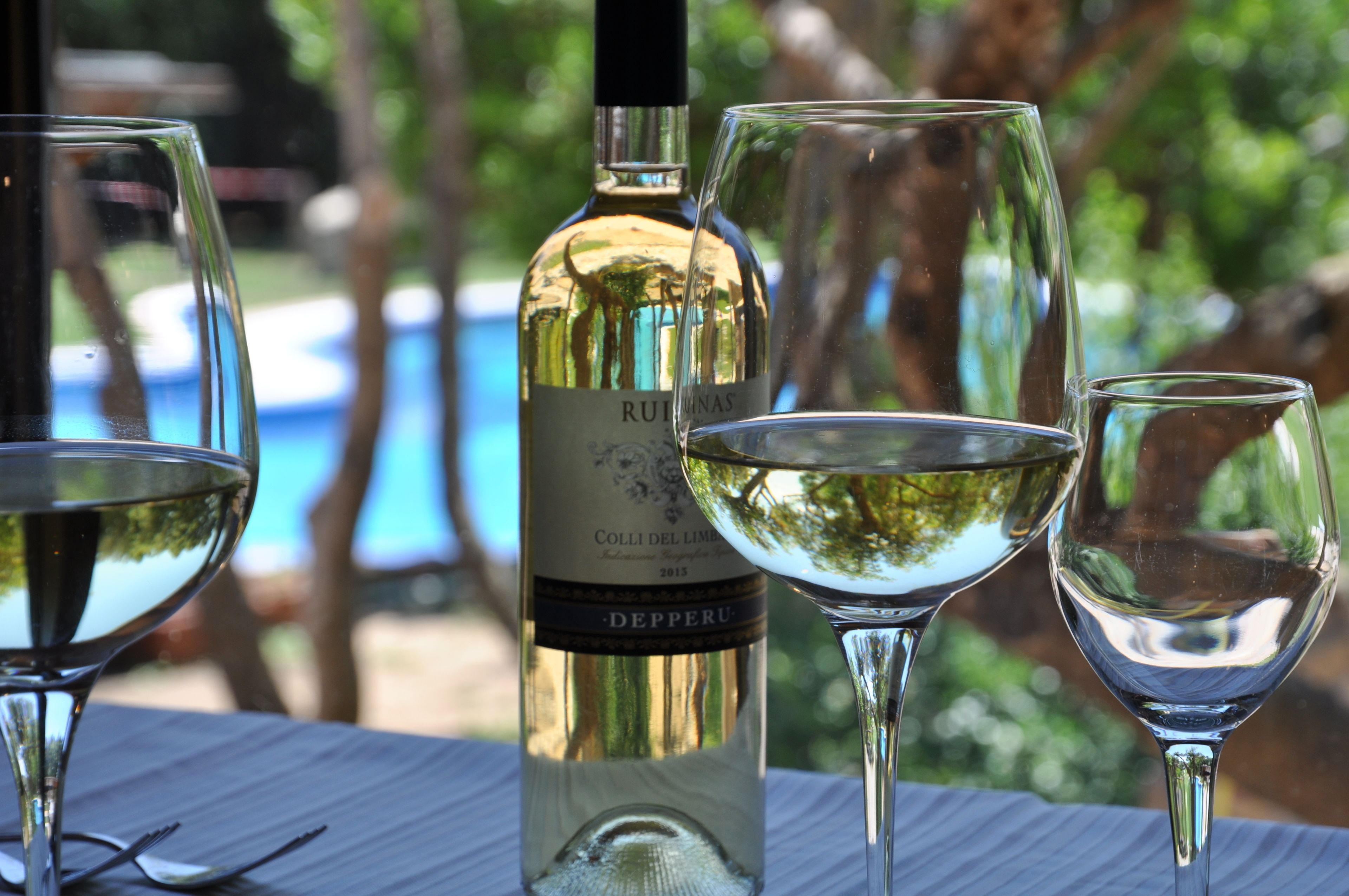 A bottle of wine and three glasses on a table with a pool in the background.