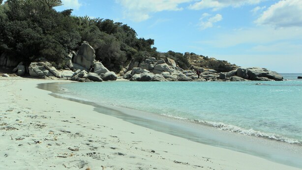 Een zandstrand met helder turquoise water, rotsformaties en weelderige begroeiing.