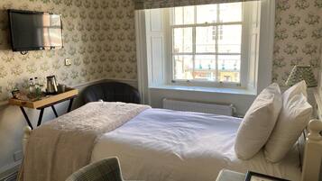 Un dormitorio ordenado con cama, una silla, un escritorio con lámpara y una ventana con vistas a edificios.