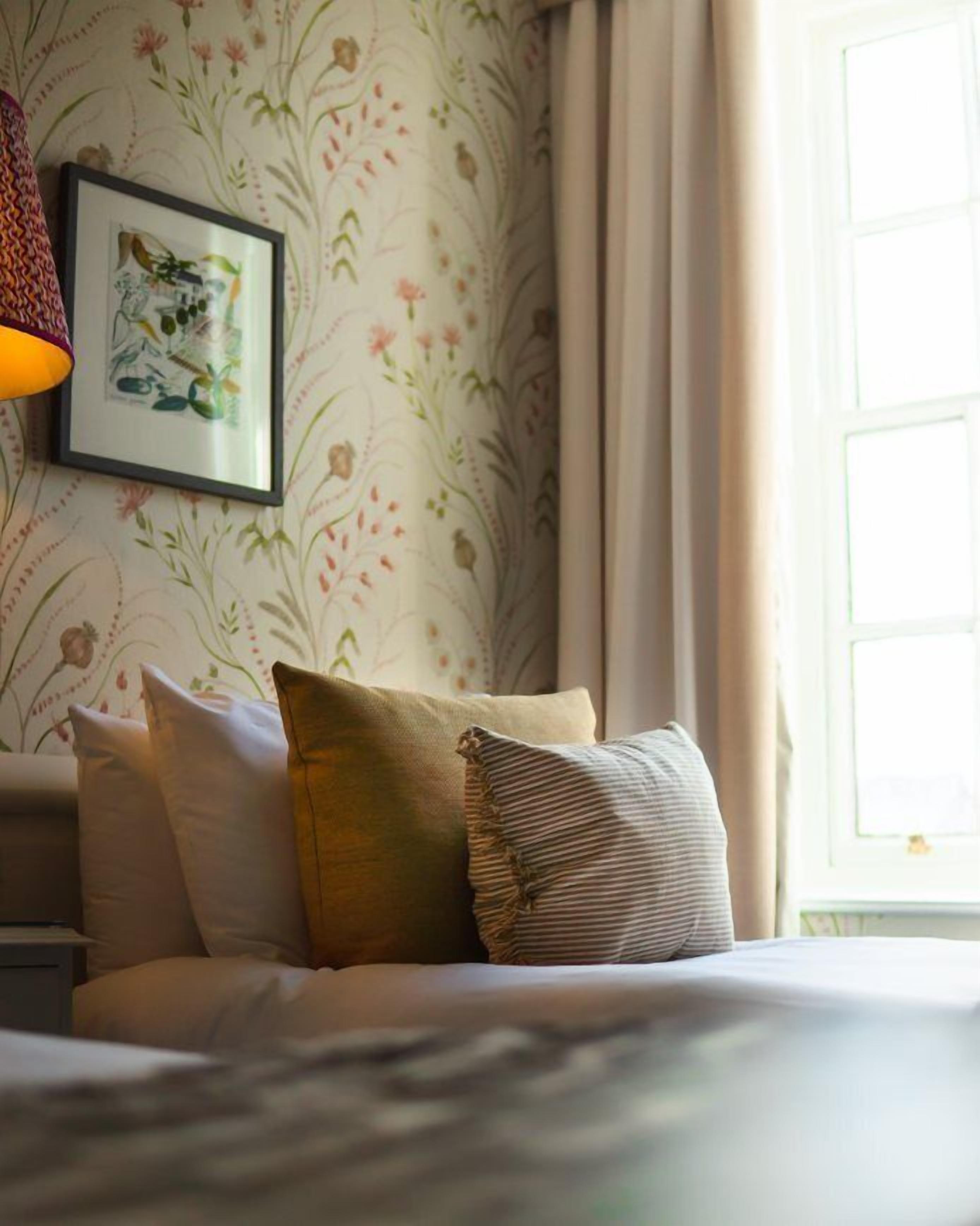 A bedroom with floral wallpaper, a framed botanical print on the wall, and a window with curtains.