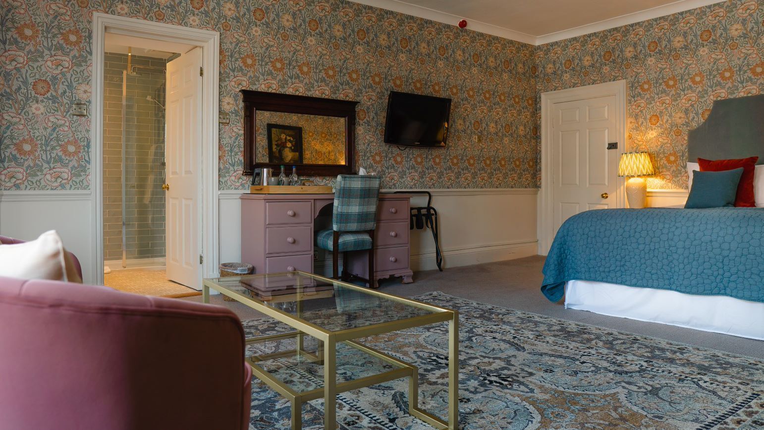 Une chambre avec un papier peint à motifs floraux, un lit, un bureau avec une chaise, un téléviseur et une salle de bain visible à travers une porte ouverte.