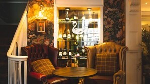 Un rincón acogedor con dos sillones de cuero y una mesita redonda, una escalera con un fondo de papel tapiz floral, y una zona de bar con botellas y una decoración de cisne.