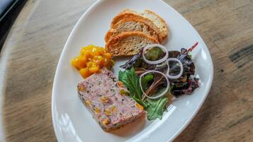 Un plato blanco con rebanadas de pan, un pastel de carne, pepinillos y una ensalada sobre una mesa de madera.
