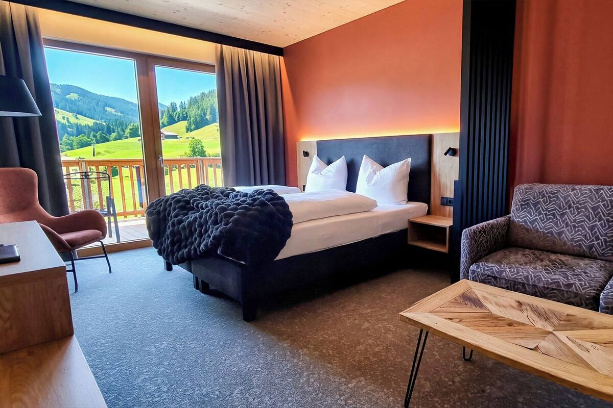 A hotel room with a bed, a sofa, a desk, and a view of a green landscape.