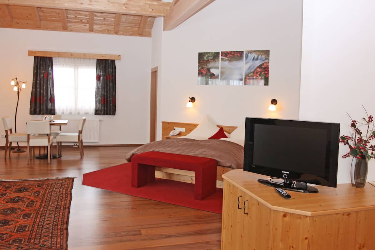 A modern hotel room with a wooden ceiling, a large bed with red bedding, a TV on a wooden cabinet, and a dining area with a table and chairs.