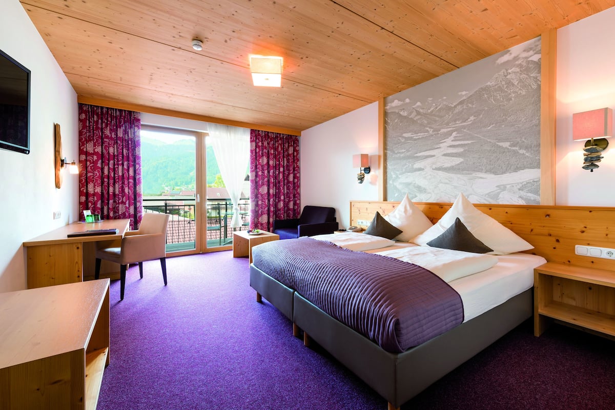 A hotel room with a bed, desk, chair, and a balcony with a mountain view.