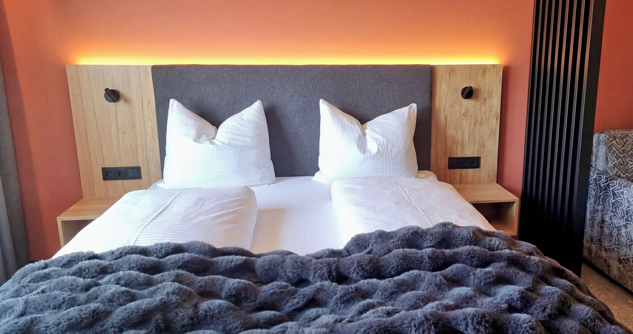 A double bed with white linens and a grey headboard, flanked by wooden nightstands with black speakers.