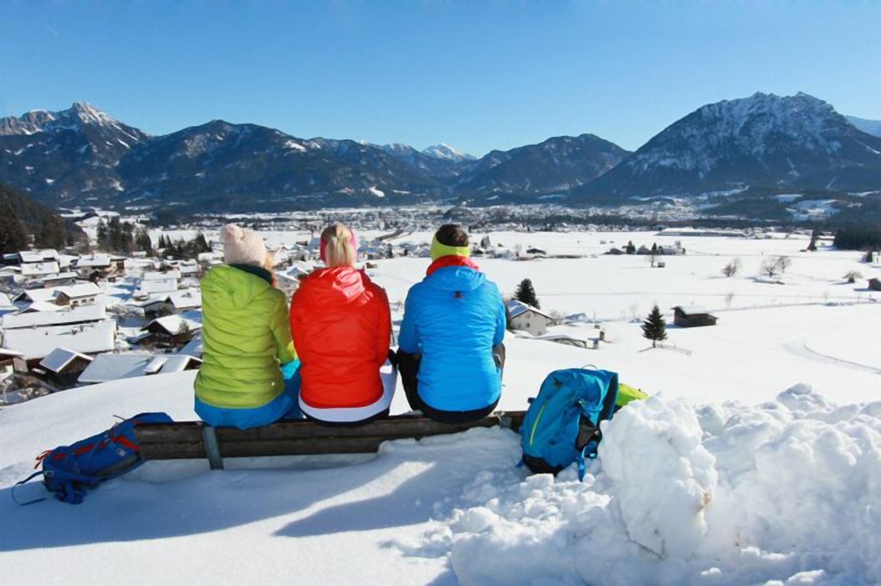 Tres personas sentadas en un banco de madera con vistas a un paisaje nevado y montañas al fondo.