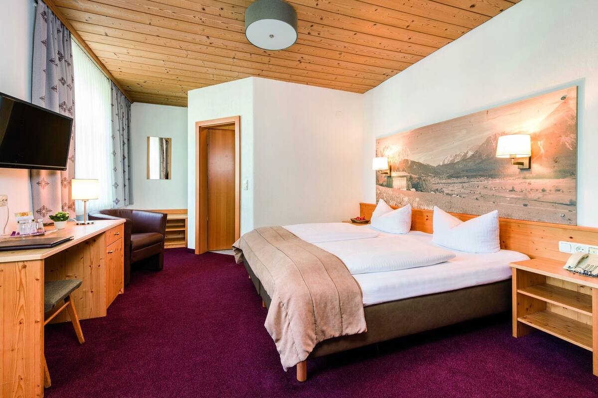 A hotel room with a bed, desk, chair, TV, and a painting on the wall.