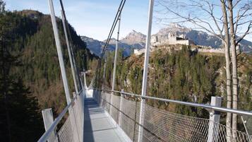 Un puente colgante con barandales y pasarela de metal, rodeado de montañas boscosas y una estructura que parece un castillo.