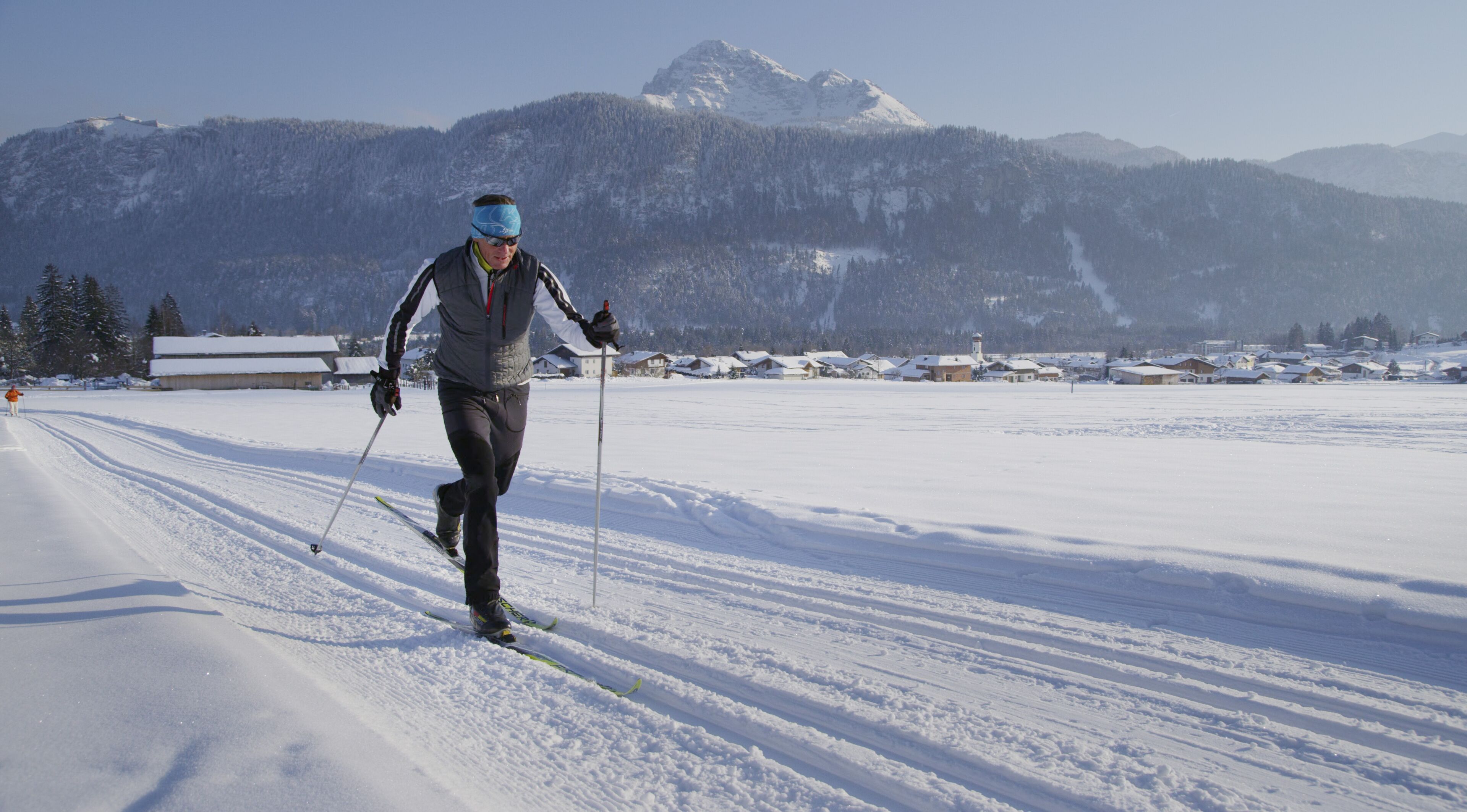 A person cross-country skiing on a snow-covered path with a mountainous backdrop.