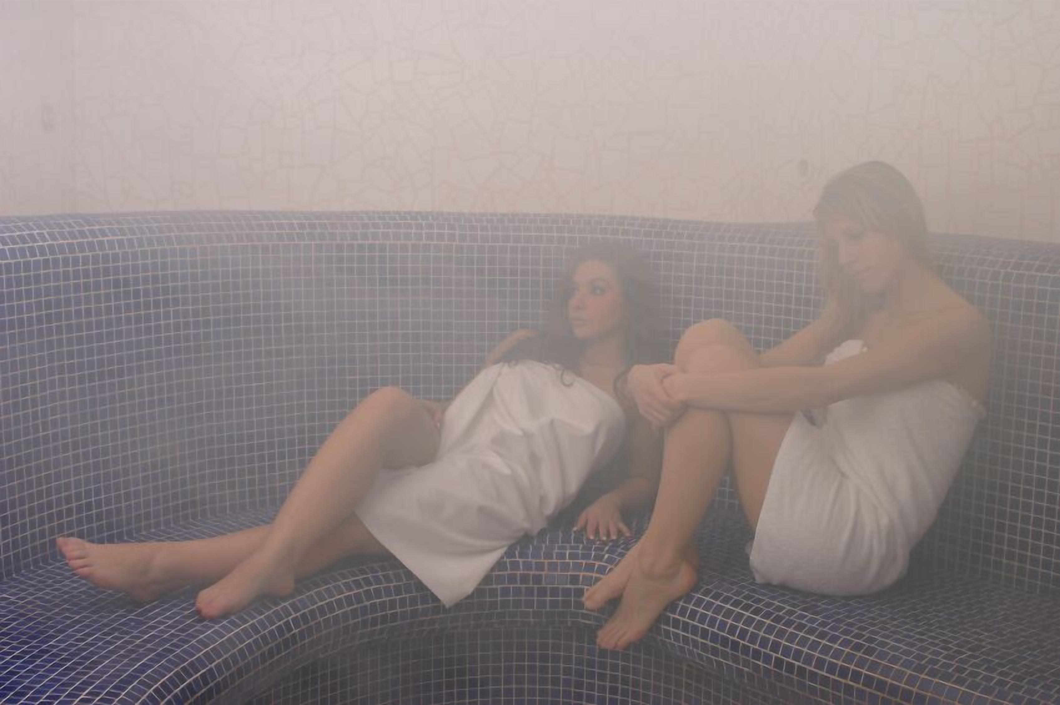 Duas pessoas sentadas em um banco de azulejos em um espaço com sauna.