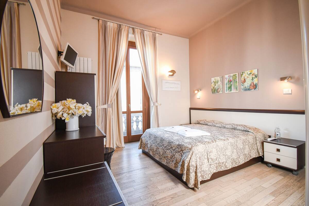 A bedroom with a bed, bedside table, nightstand, dresser, mirror, and a window with curtains.