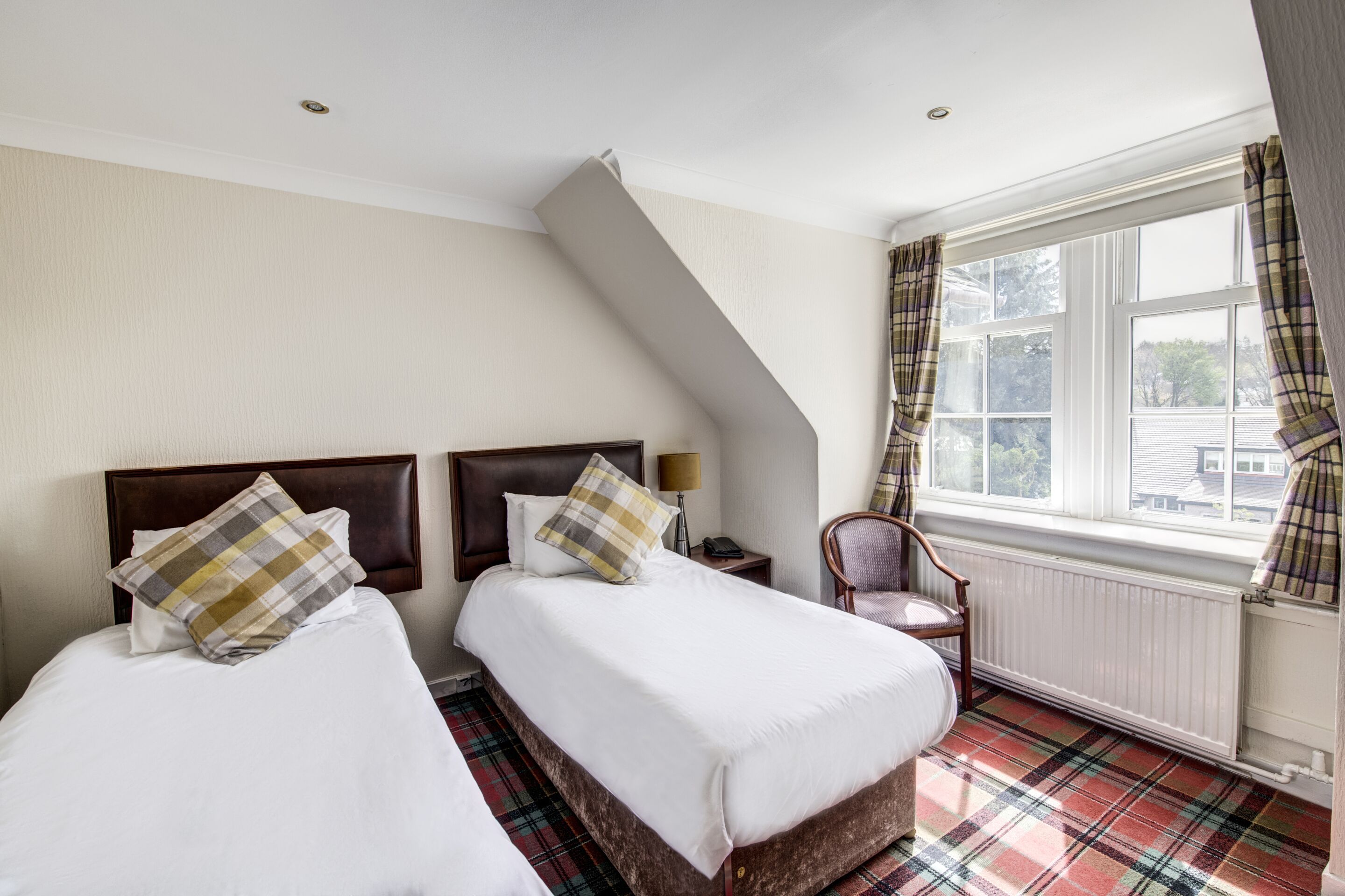 A hotel room with two beds, a chair, a window with curtains, and a radiator.