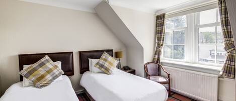 A hotel room with two beds, a chair, a window with curtains, and a radiator.