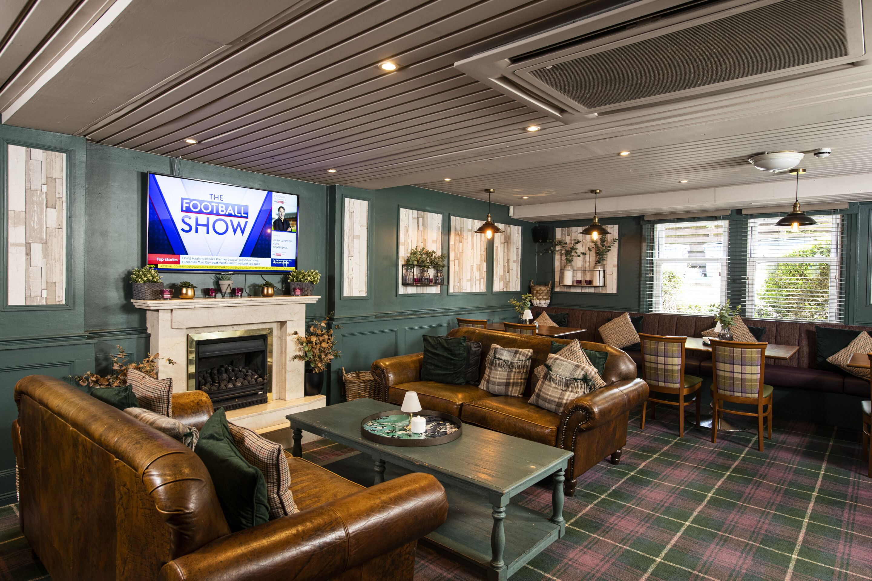 A cozy living room with a fireplace, leather sofas, a television displaying 'The Football Show', and a plaid carpet.