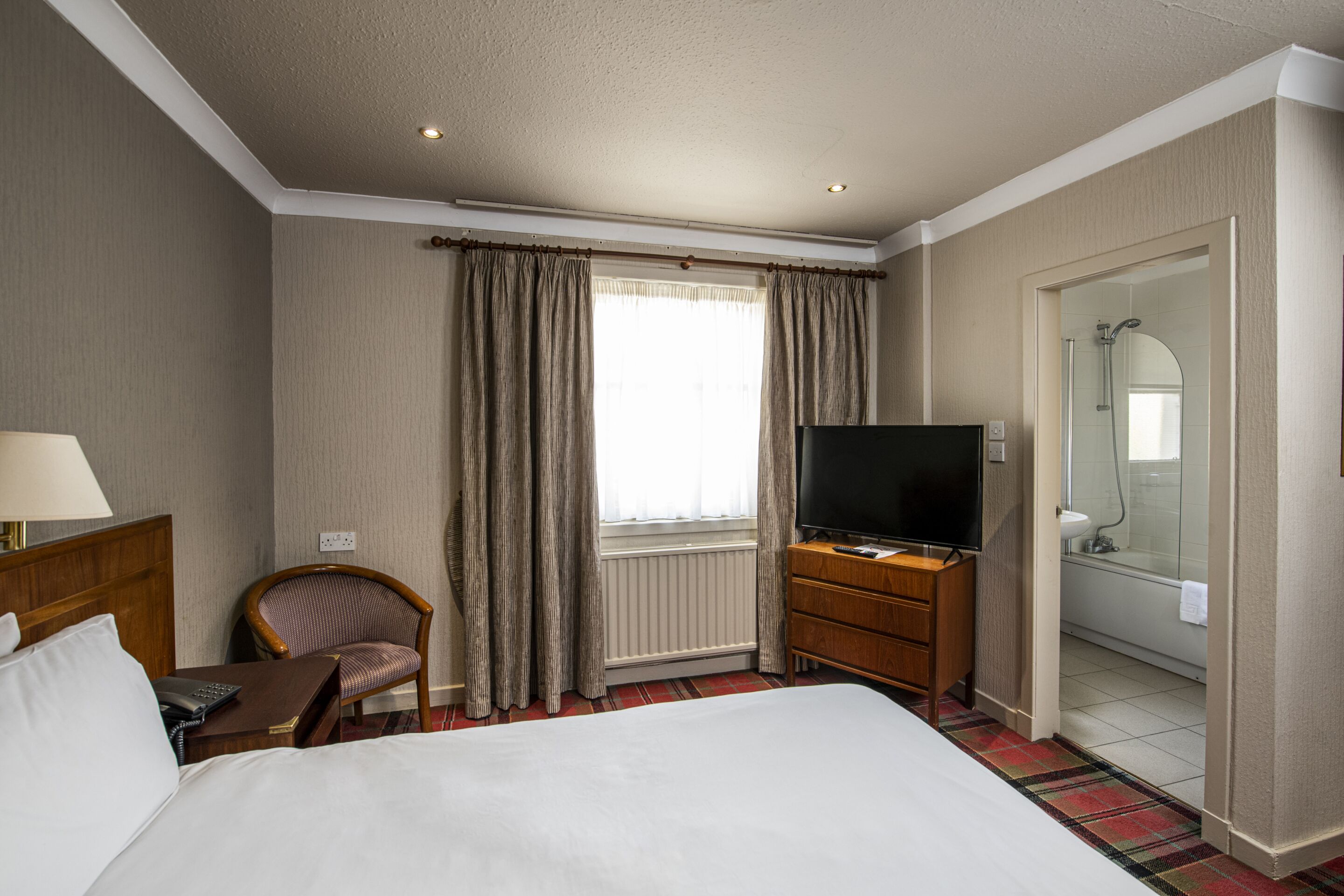 Habitación de hotel con cama, televisión, una silla, cómoda y baño visible a través de una puerta abierta.