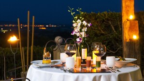 Mesa de comedor al aire libre con copas de vino, velas y una botella de vino, con un paisaje urbano de fondo.