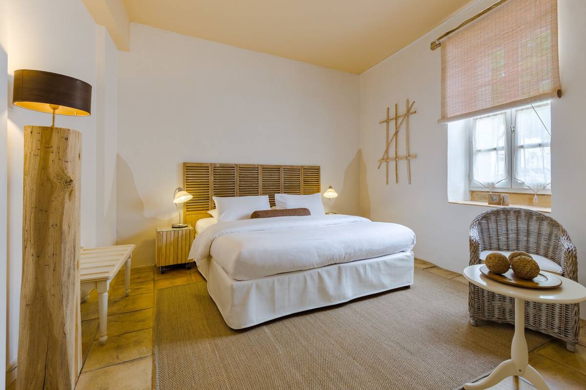 A bedroom with a large bed, bedside lamps, a wooden headboard, a wicker chair, and a small table with decorative items.