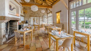 Un restaurante con paredes de piedra, vigas de madera y ventanas amplias. Las mesas están dispuestas con manteles blancos y velas.