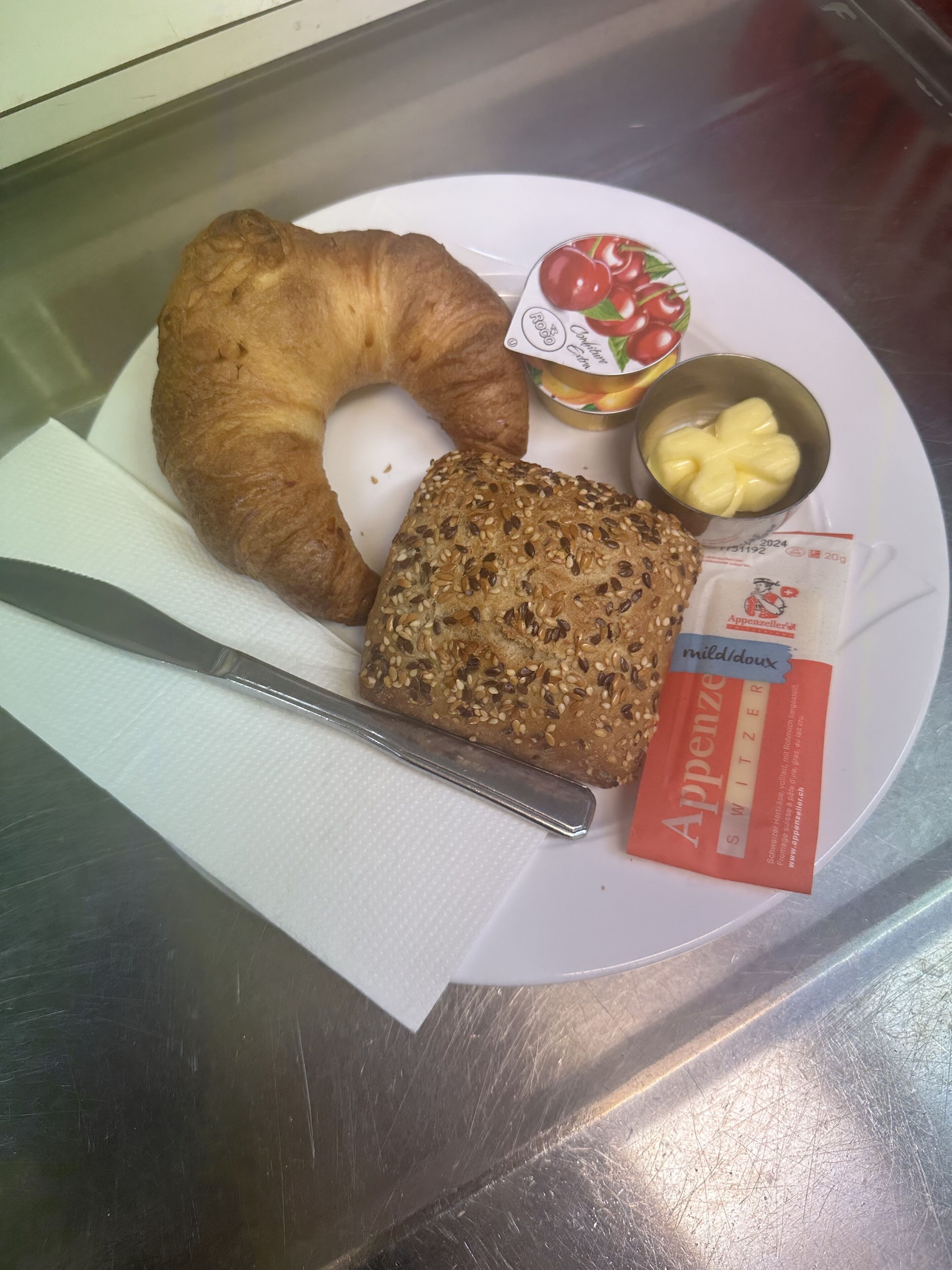 A breakfast plate with a croissant, a slice of multigrain bread, a small container of butter, a cup of yogurt, and a packet of Appenzeller cheese.