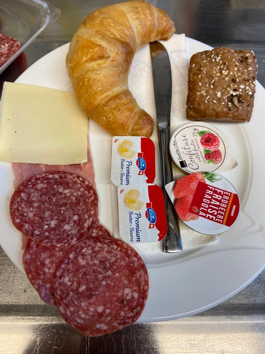 Un plato con un croissant, queso, salami, pan, mantequilla y mermelada.
