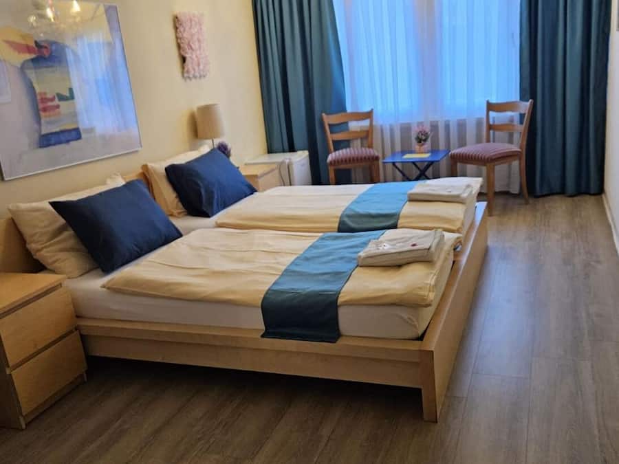 Una cama doble con cubrecama azul, una mesita de noche de madera, una silla, una pequeña mesa y una araña.