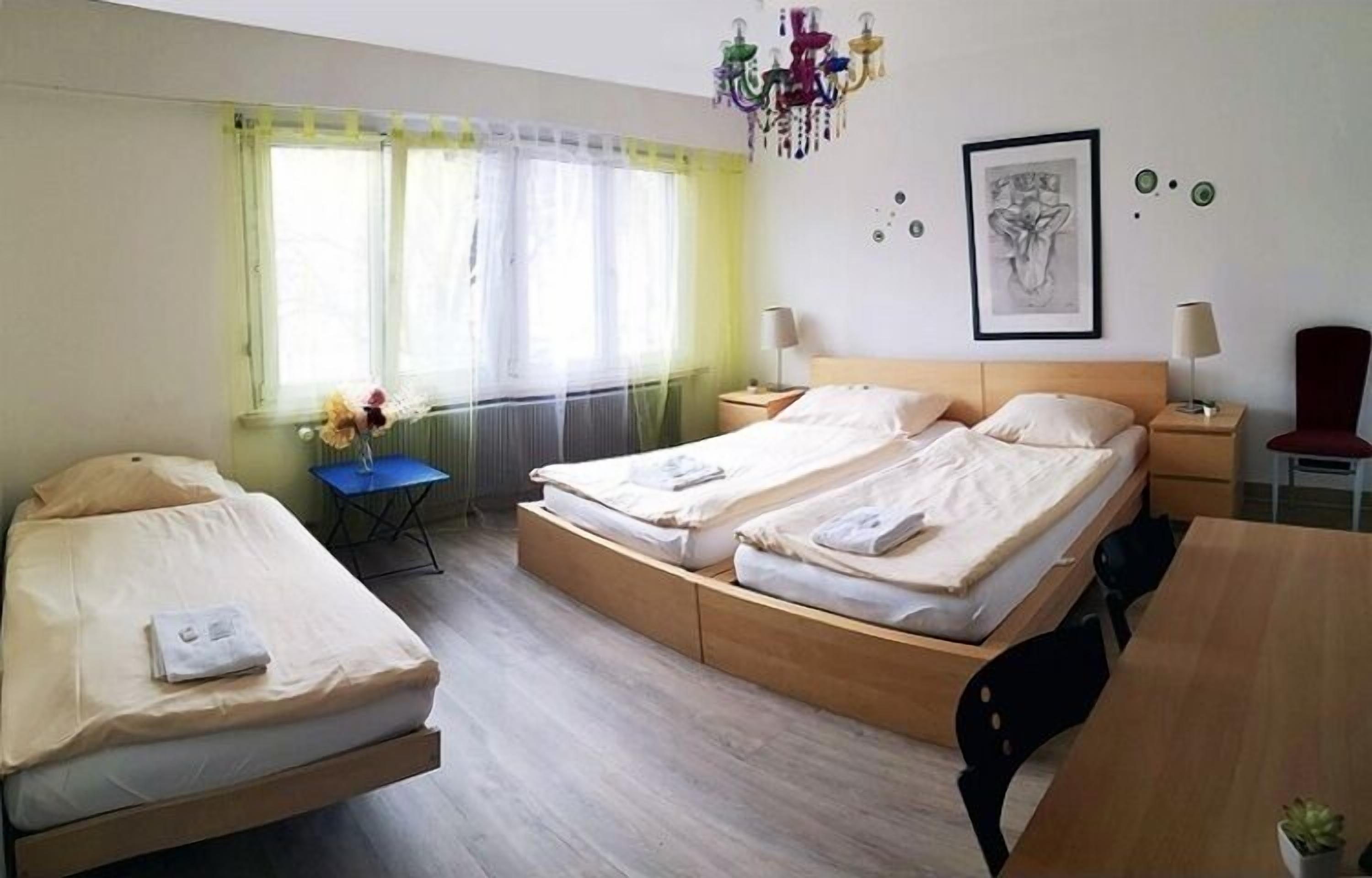 Quarto de hotel com duas camas de solteiro, uma escrivaninha de madeira, uma cadeira, uma mesinha e uma janela com cortinas.