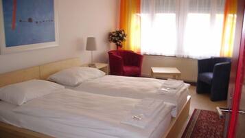 Habitación de hotel con dos camas, una silla roja, una mesita y un ventanal con cortinas.