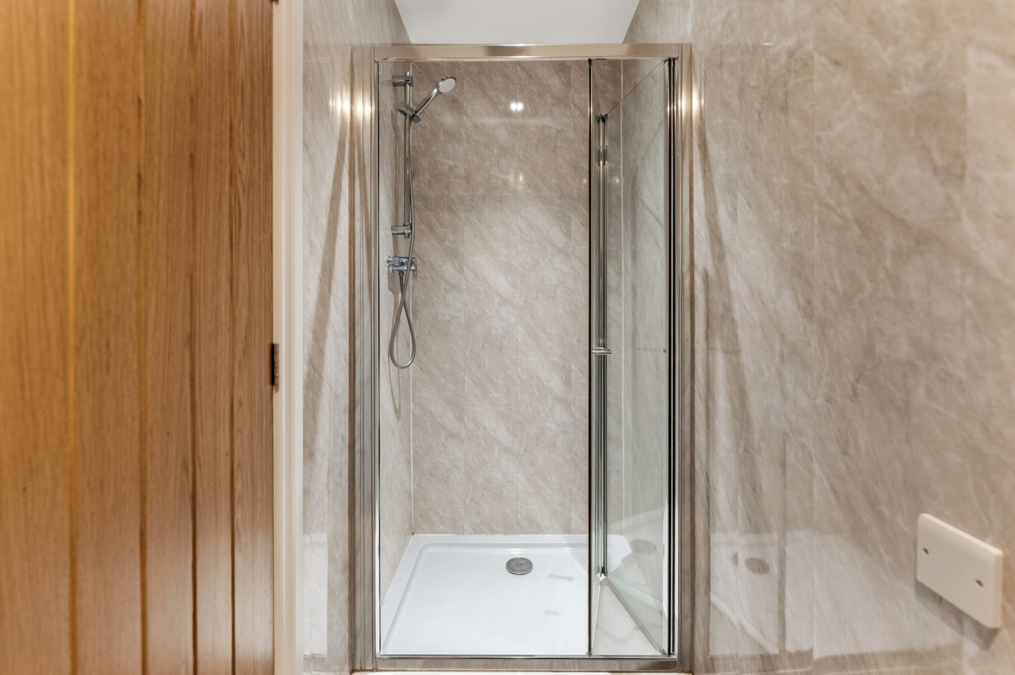 A glass-enclosed shower with a white floor, a wooden door, and a marble wall.