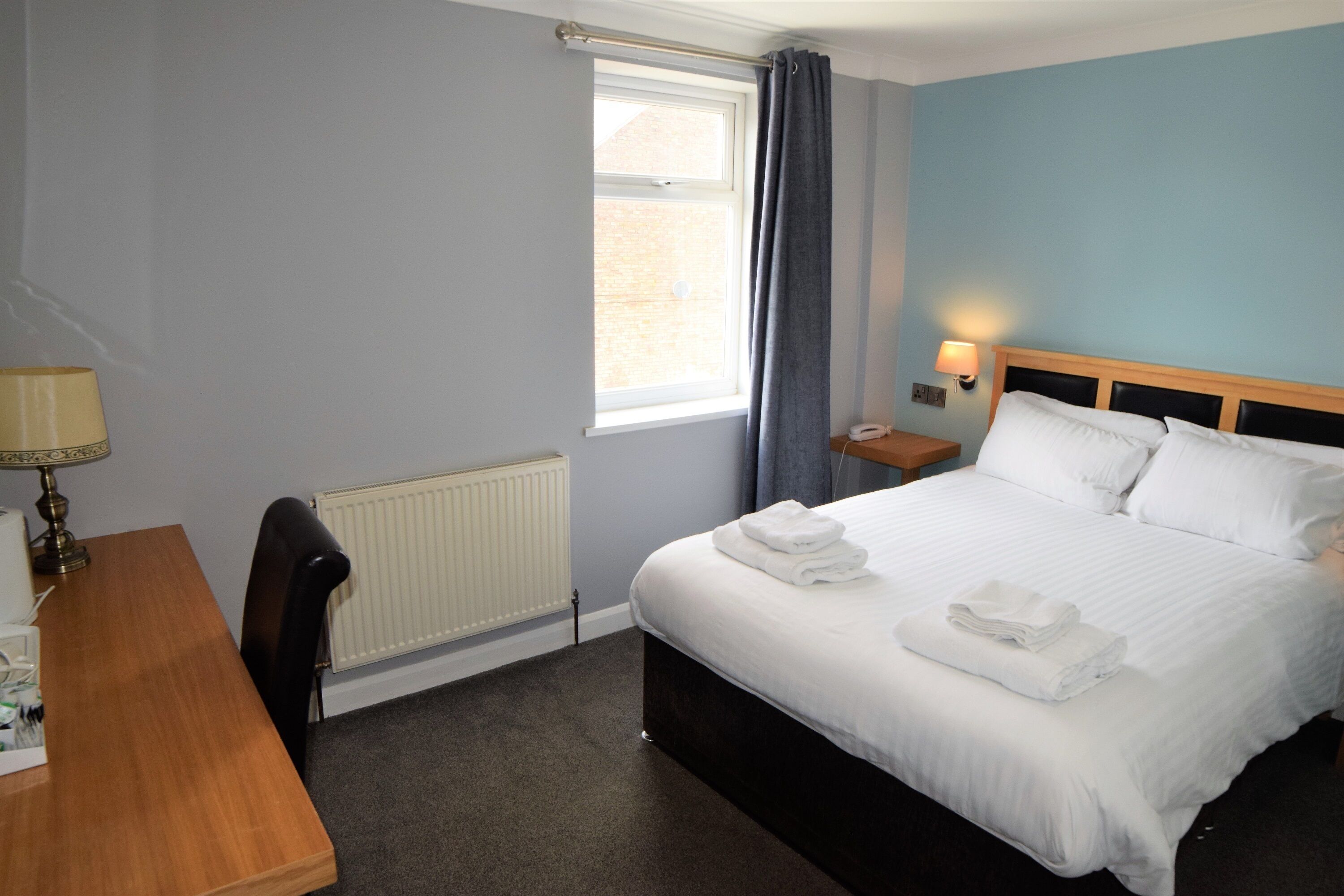 A hotel room with a bed, a desk, a chair, a window with curtains, a nightstand with a lamp, and a radiator.