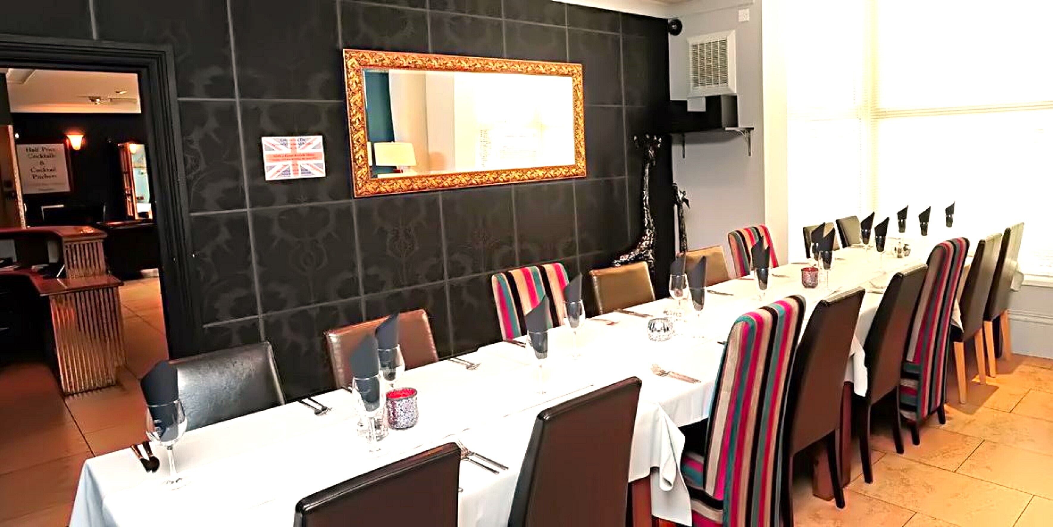 Un comedor con una larga mesa dispuesta para una comida, sillas con cojines a rayas, un espejo en la pared y un cartel que anuncia un menú a mitad de precio.