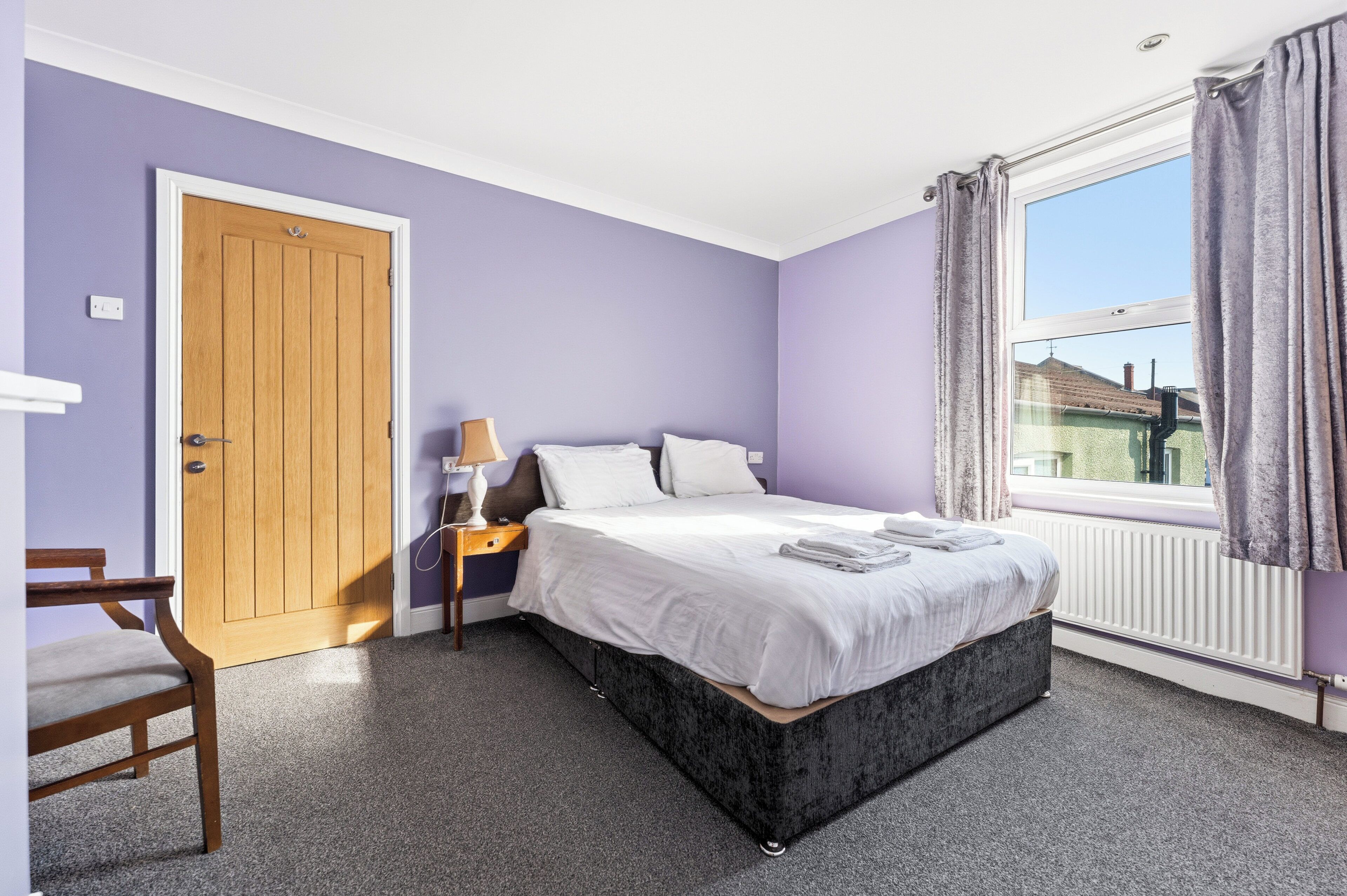 A bedroom with a bed, a nightstand, a lamp, a chair, a window with curtains, and a door.
