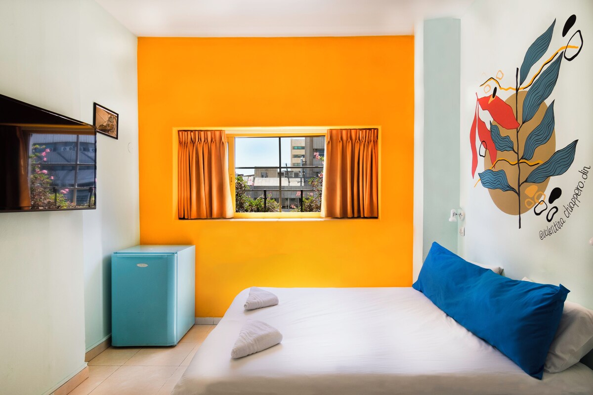 A bedroom with a bed, a small blue refrigerator, a window with curtains, and a wall art piece.